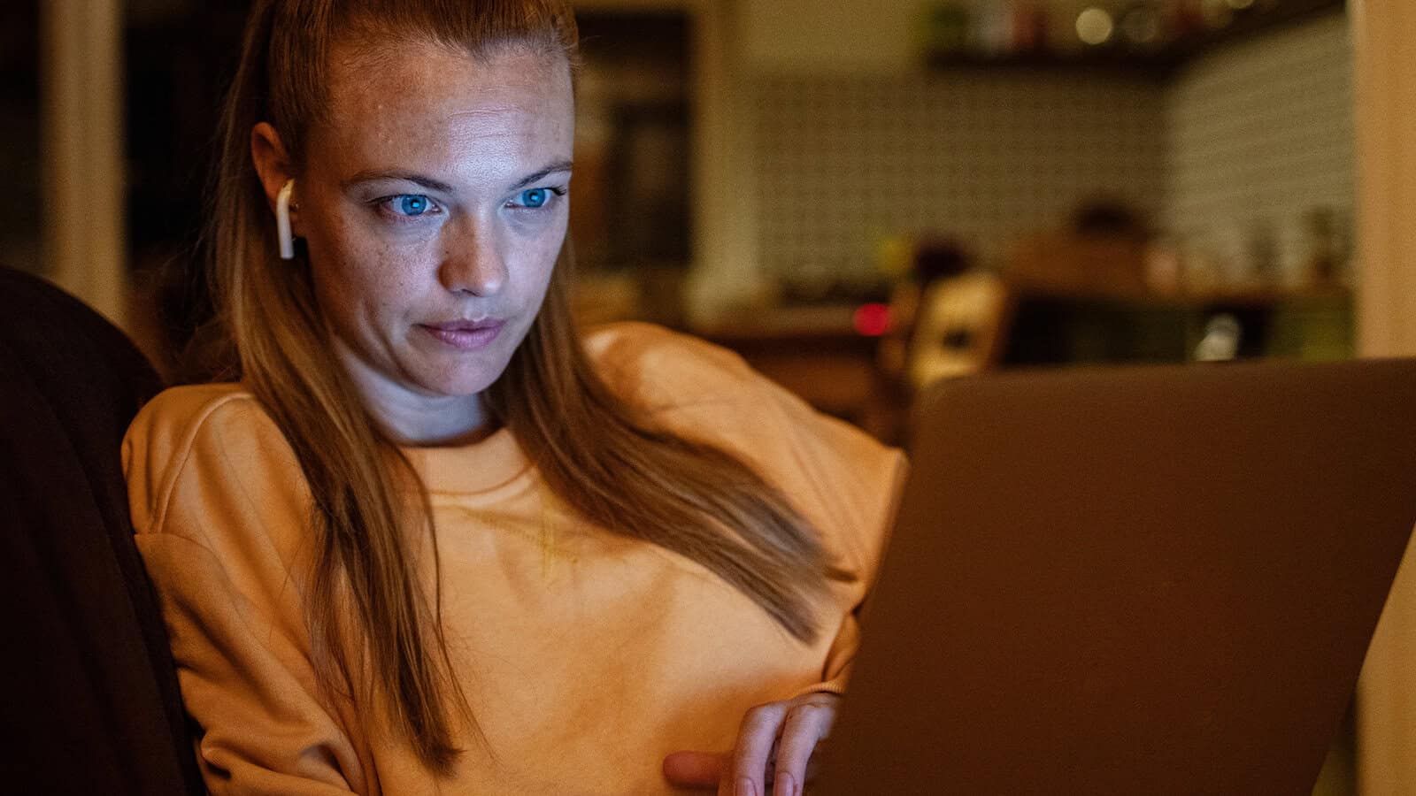 A woman with long red hair, wearing a peach sweater and wireless earbuds, sits indoors and looks at a laptop screen with a focused expression.