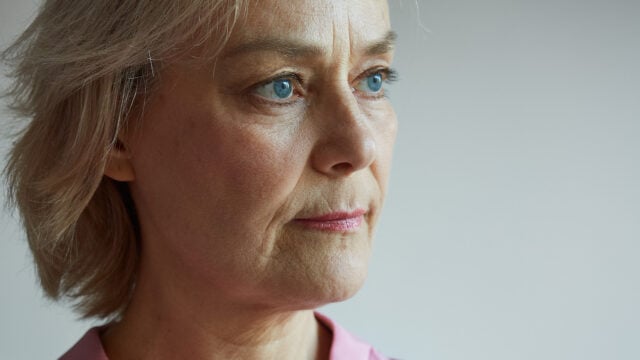 A close-up of a middle-aged woman with short blonde hair and blue eyes, gazing thoughtfully into the distance. She is wearing a light pink top and appears to be deep in thought.