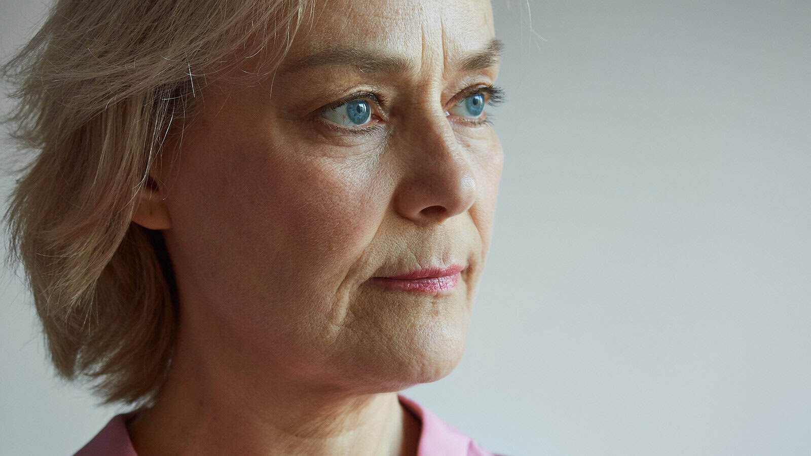 A close-up of a middle-aged woman with short blonde hair and blue eyes, gazing thoughtfully into the distance. She is wearing a light pink top and appears to be deep in thought.