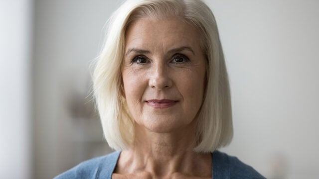 A woman with short, straight, white-blonde hair and light skin smiles gently, wearing a blue top. The background is softly blurred, focusing on her face.