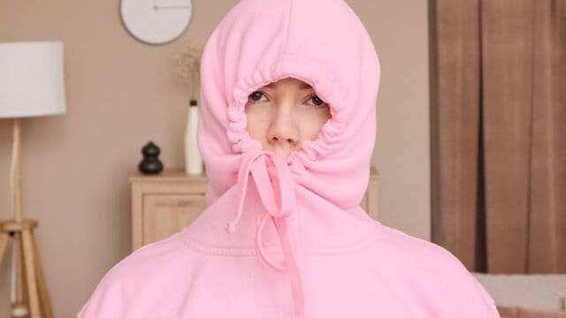 A person wears a pink hoodie with the hood tightly pulled around their face, exposing only their eyes and nose. A clock, lamp, and cabinet are visible in the softly lit background.