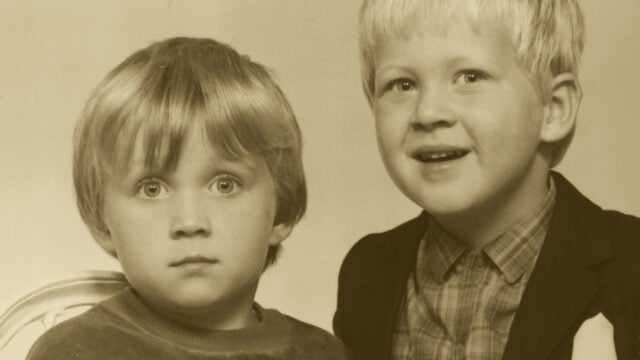 Two young children with light hair pose for a portrait. One has a neutral expression and wears a sweater, while the other smiles and wears a patterned shirt with a jacket. The photo is sepia-toned.