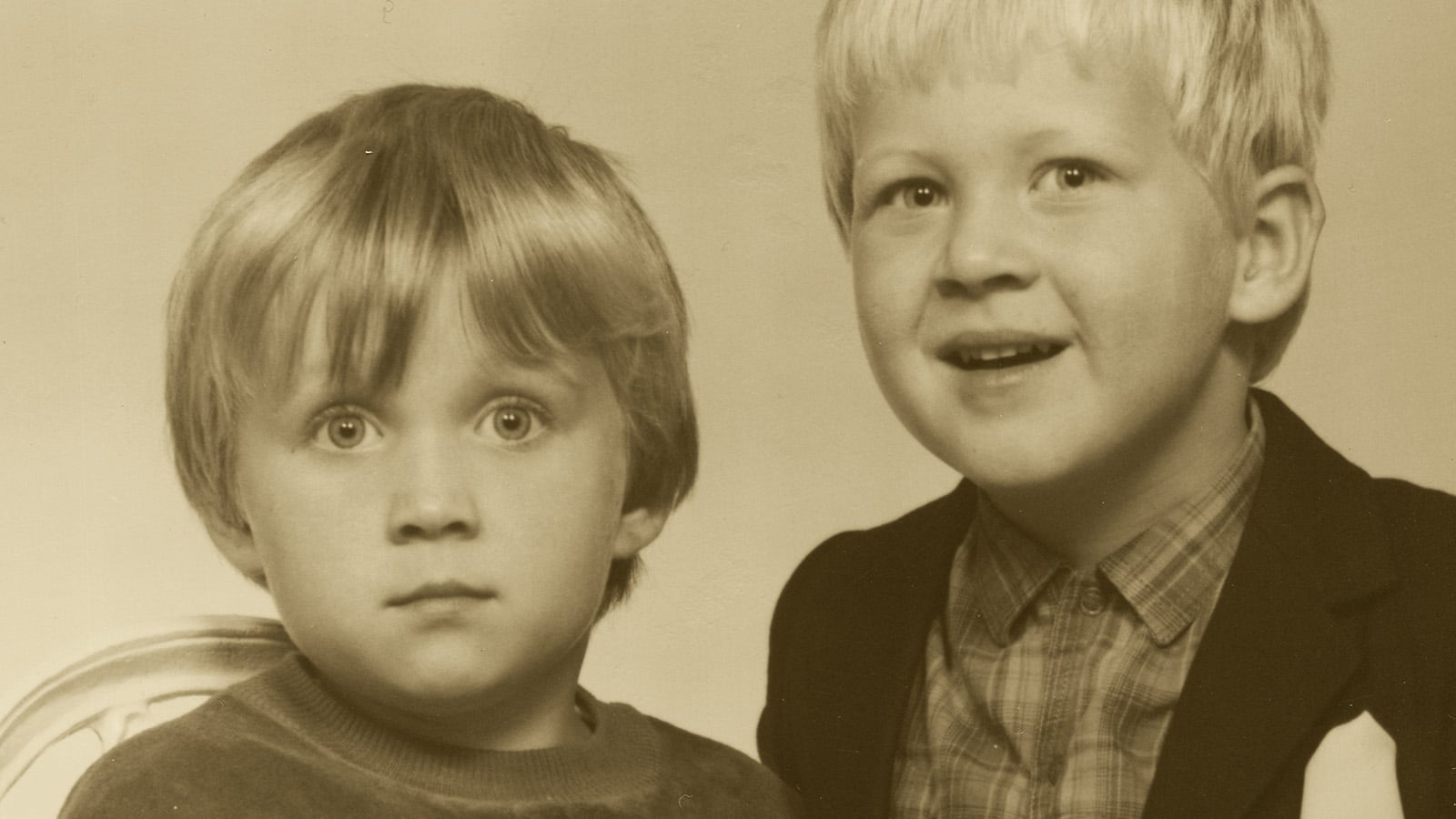 Two young children with light hair pose for a portrait. One has a neutral expression and wears a sweater, while the other smiles and wears a patterned shirt with a jacket. The photo is sepia-toned.