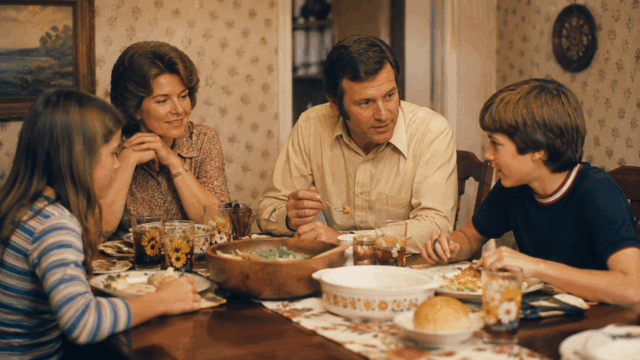 A family of four sits around a dinner table, smiling and talking while eating a meal. The table is set with food and drinks, and the room has floral wallpaper and framed art on the wall.