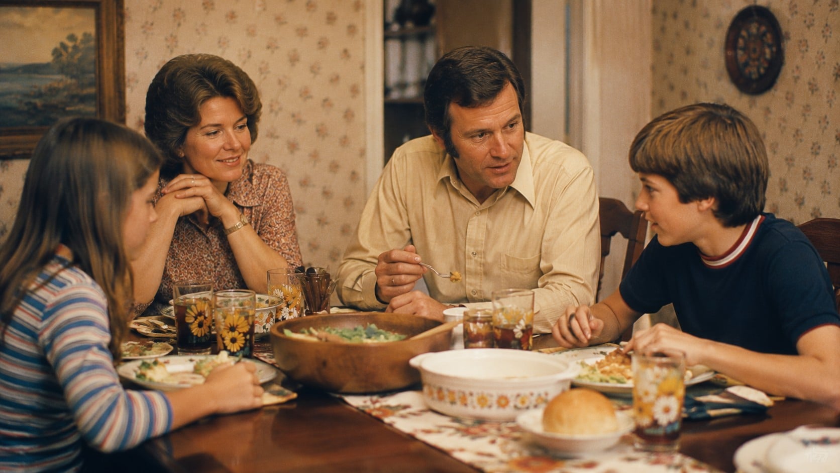 A family of four sits around a dinner table, smiling and talking while eating a meal. The table is set with food and drinks, and the room has floral wallpaper and framed art on the wall.