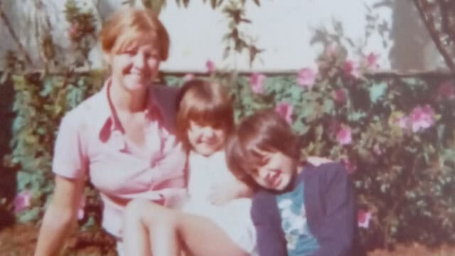 A smiling woman sits outdoors with two young children in her lap, surrounded by blooming pink flowers and greenery on a sunny day.
