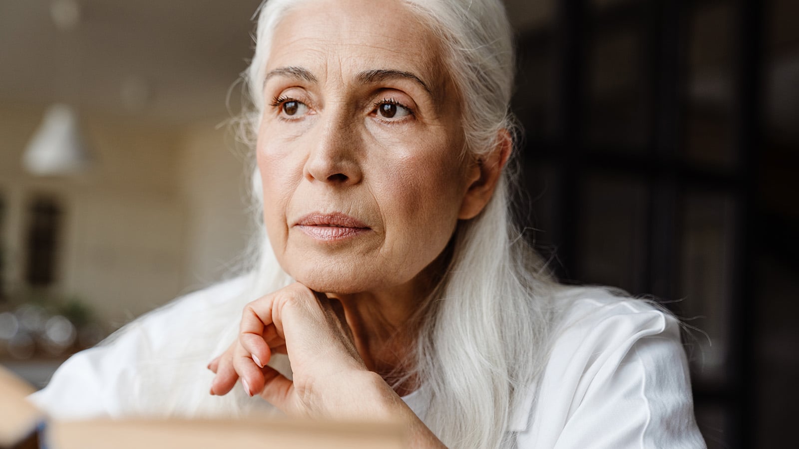 An older woman with long gray hair and a thoughtful expression rests her chin on her hand while looking into the distance indoors.