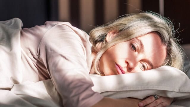 A woman with blonde hair lies on her side in bed, resting her head on a pillow and looking thoughtfully ahead. Sunlight casts shadows across her face, giving a calm yet pensive mood.