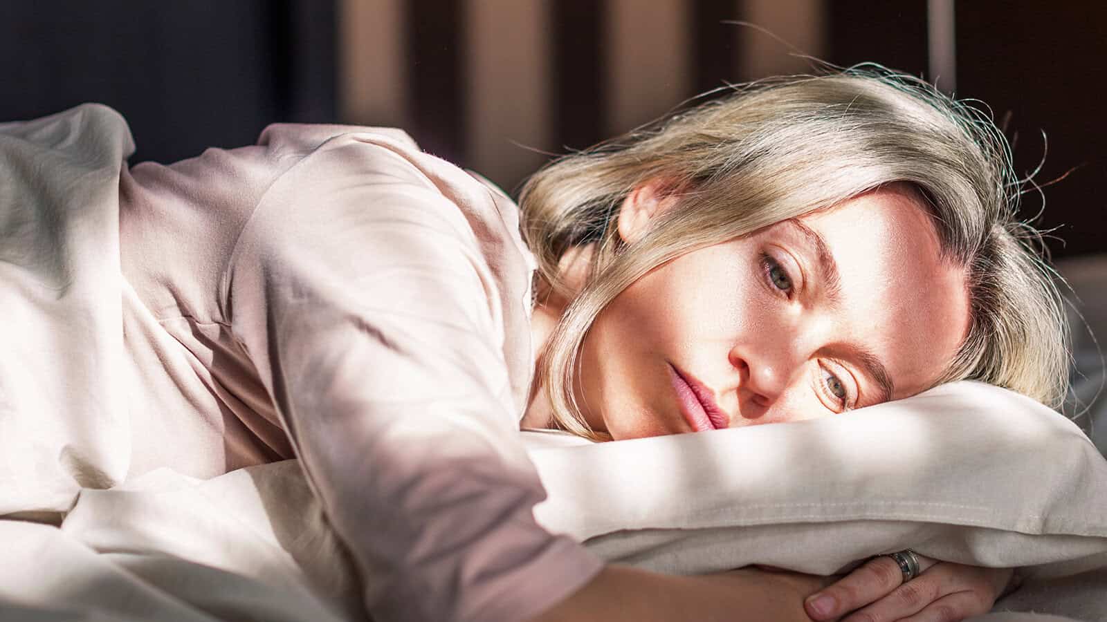 A woman with blonde hair lies on her side in bed, resting her head on a pillow and looking thoughtfully ahead. Sunlight casts shadows across her face, giving a calm yet pensive mood.