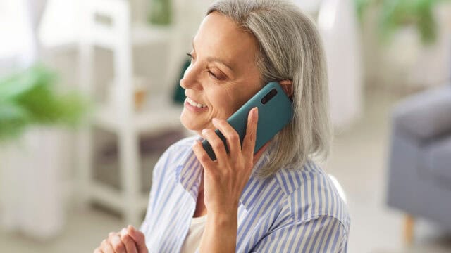 Smiling older woman with gray hair talks on a smartphone while sitting indoors, wearing a striped blue and white shirt.