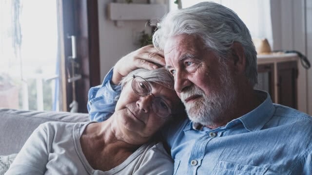 An elderly couple sits closely together on a couch, with the man gently resting his hand on the womans head. They both appear calm and content, sharing a quiet, intimate moment in a softly lit room.
