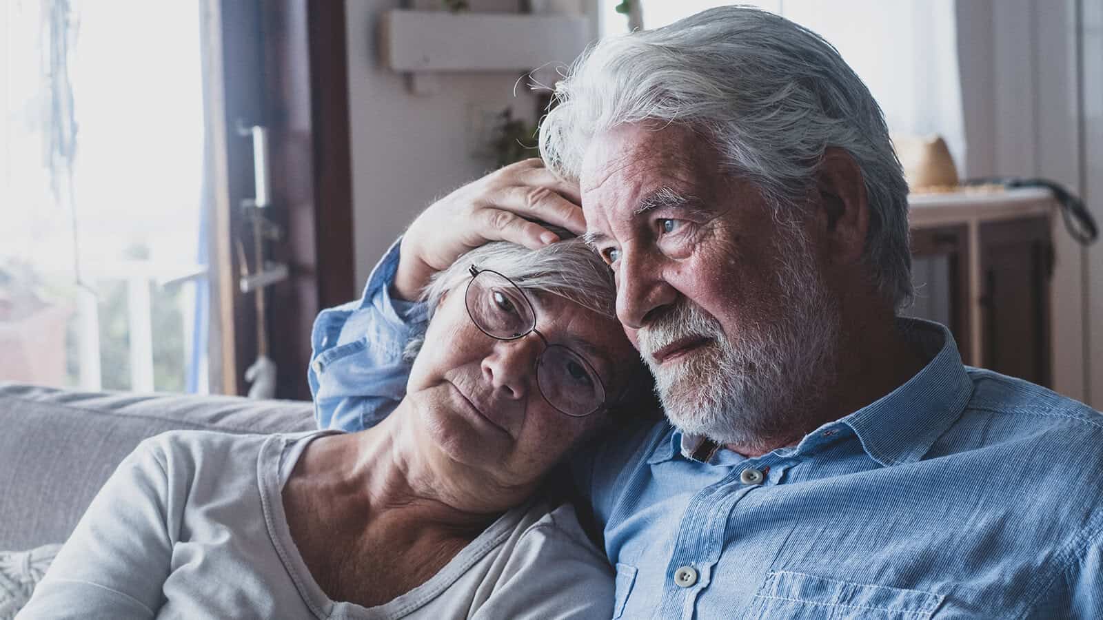 An elderly couple sits closely together on a couch, with the man gently resting his hand on the womans head. They both appear calm and content, sharing a quiet, intimate moment in a softly lit room.