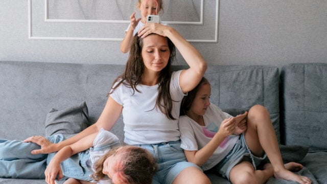 A tired woman sits on a couch with three children climbing on and around her. One child plays with her hair, another leans on her lap, and a third looks at a smartphone.