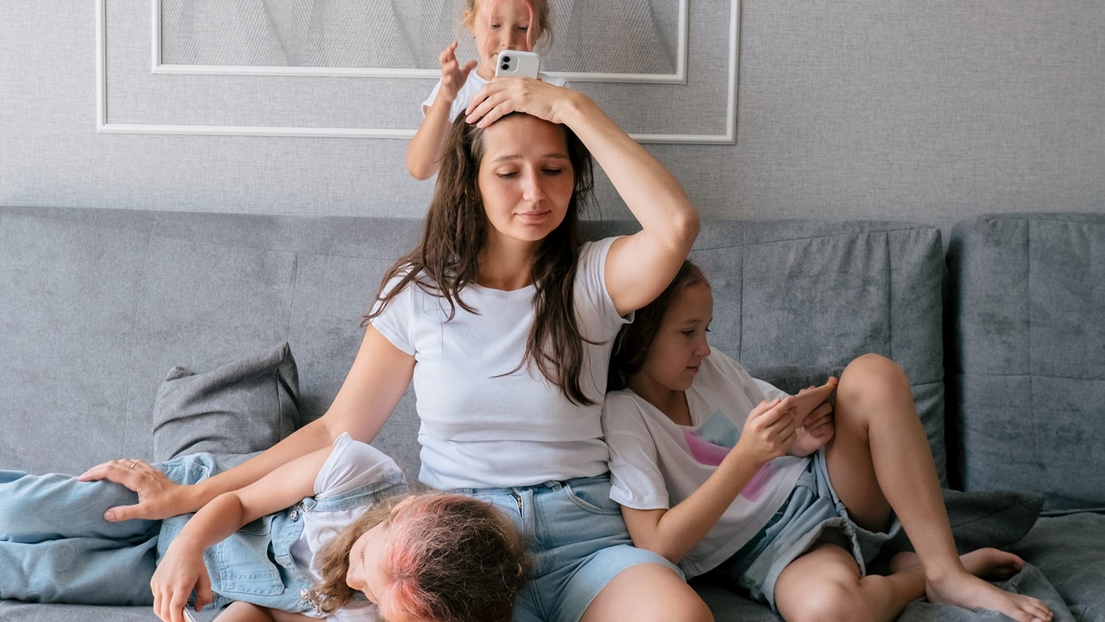 A tired woman sits on a couch with three children climbing on and around her. One child plays with her hair, another leans on her lap, and a third looks at a smartphone.