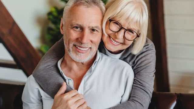 Smiling older woman with glasses hugs a smiling older man from behind while sitting indoors. Both wear casual clothes and look happy and relaxed.