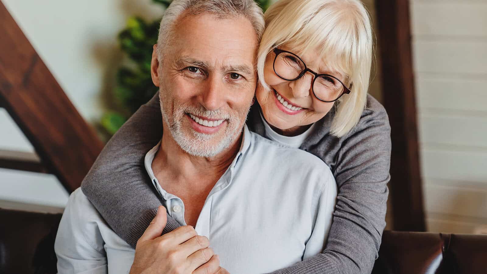 Smiling older woman with glasses hugs a smiling older man from behind while sitting indoors. Both wear casual clothes and look happy and relaxed.