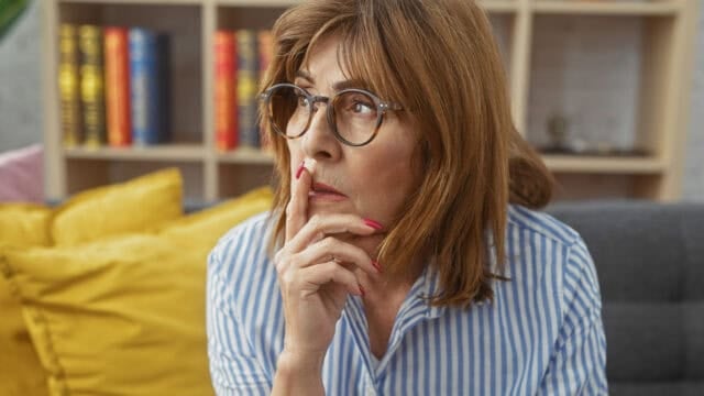 A woman with glasses and shoulder-length brown hair sits on a couch, looking thoughtful with her finger on her lips. She wears a blue and white striped shirt. Yellow and gray pillows are behind her, and bookshelves are in the background.