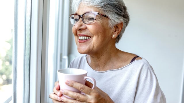Smiling older woman with short gray hair and glasses holds a white mug while looking out a window, wearing a light sweater in a bright room.