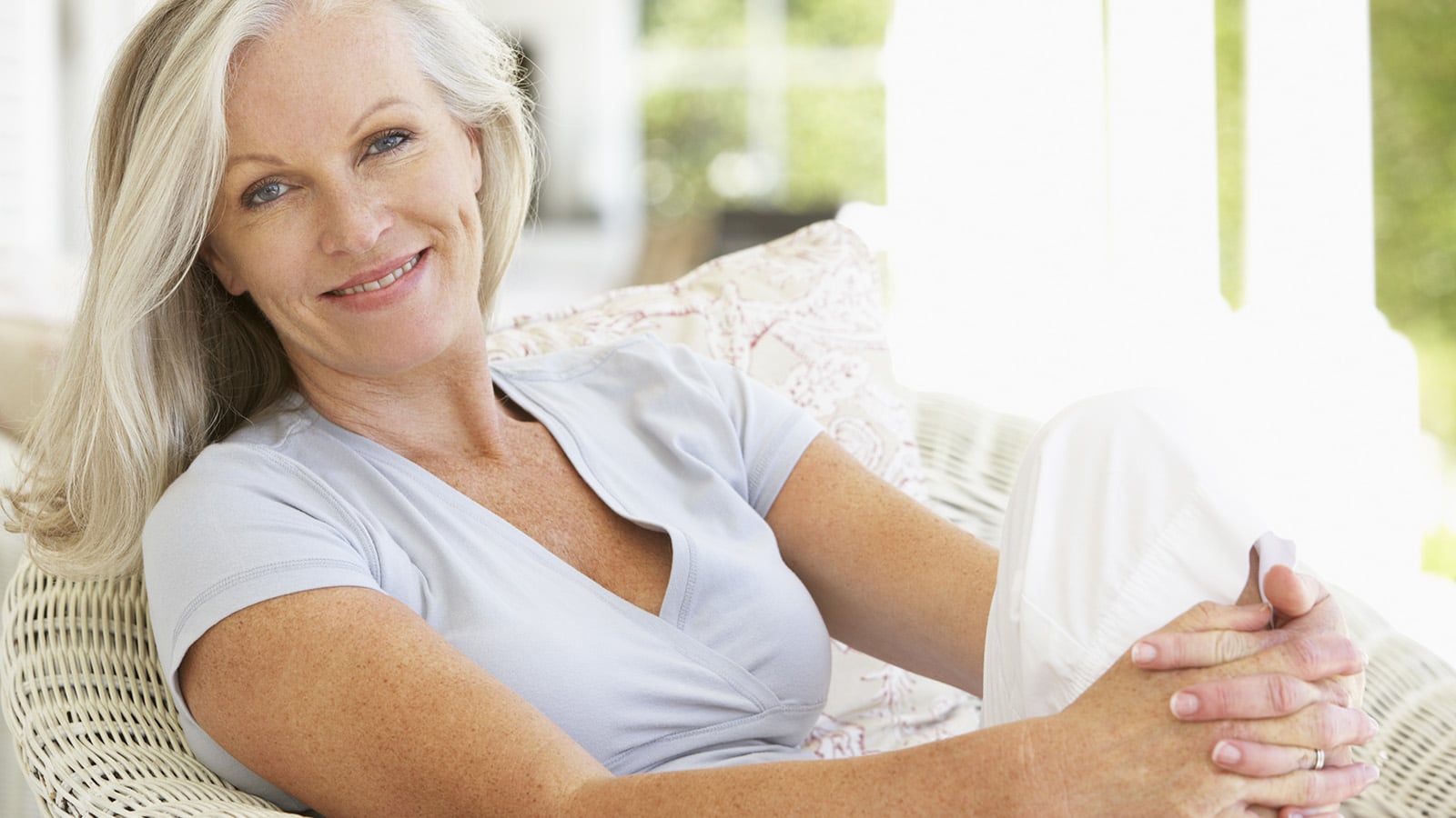 Woman with long gray hair wearing a light blue shirt and white pants, sitting on a wicker chair, smiling and looking at the camera in a bright, airy room.