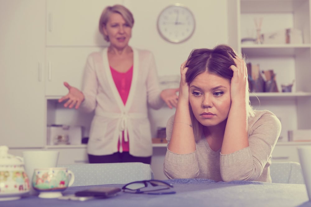 A young woman sits at a table looking upset, holding her head, while an older woman stands behind her gesturing with open arms, appearing frustrated. Their interaction highlights the challenging behaviors that can arise between adult children and their parents. A clock and shelves are visible in the background.