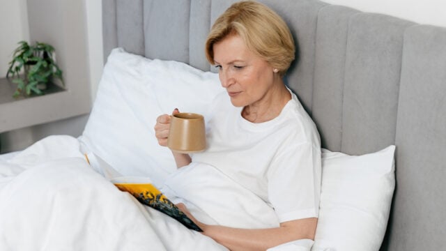 An older woman in her 70s with short blonde hair sits up in bed, enjoying her morning routines. She holds a mug and reads a book, dressed in a white t-shirt and covered by a white blanket. A small plant adds warmth to the background.