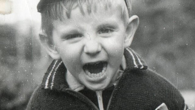 Black and white photo of a young boy with an open mouth, possibly yelling or laughing, wearing a sweater and a cap, looking directly at the camera.