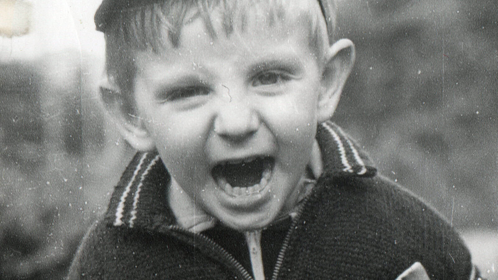 Black and white photo of a young boy with an open mouth, possibly yelling or laughing, wearing a sweater and a cap, looking directly at the camera.