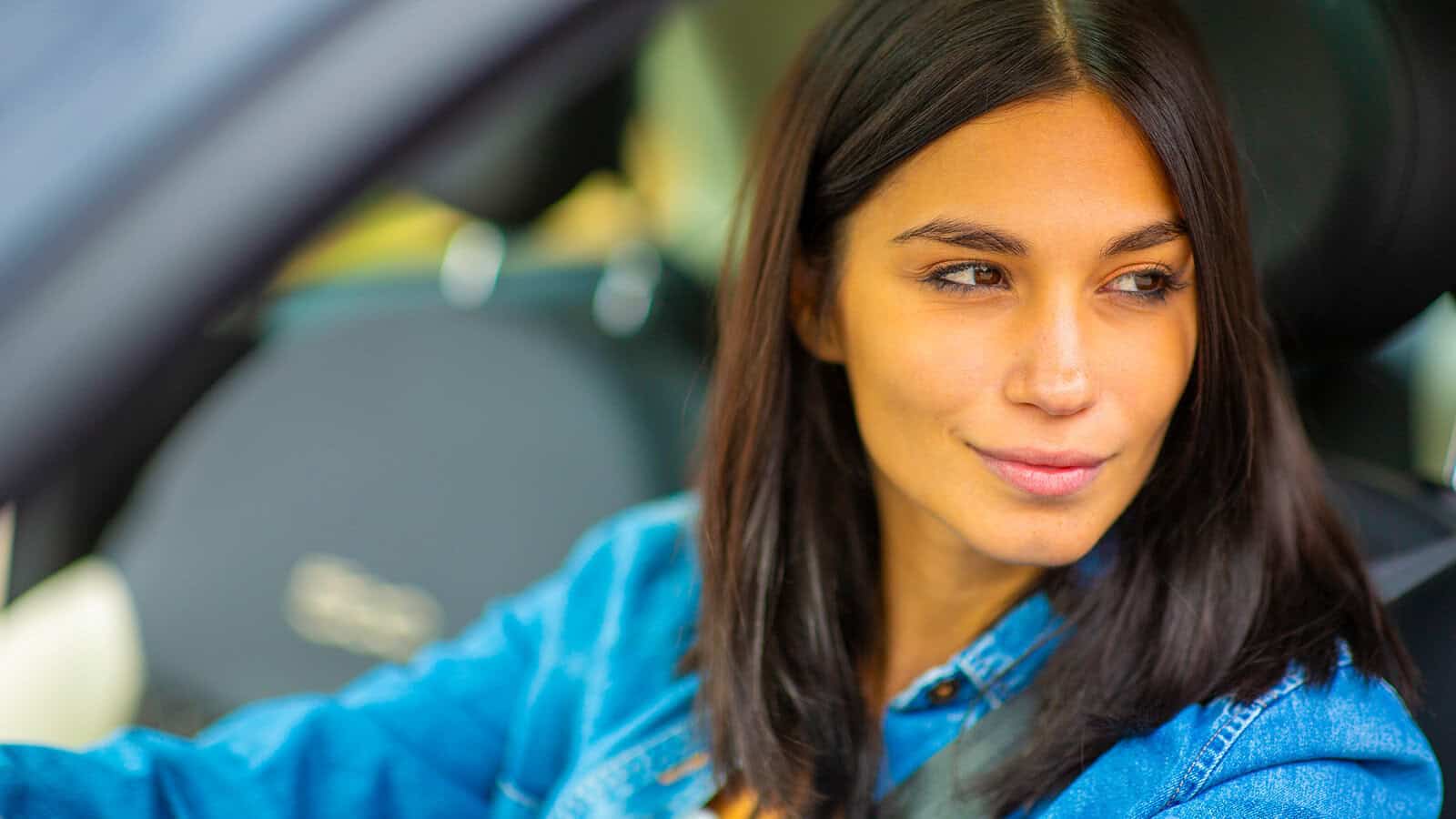 Woman with straight dark hair in a denim jacket sits in a car, looking out the window with a slight smile. The image is bright and the background is blurred.
