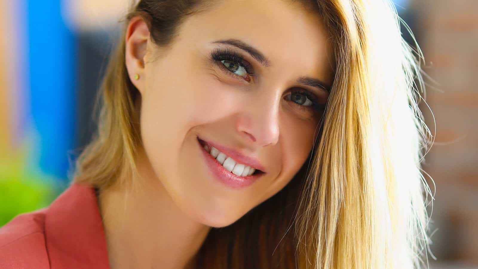 A woman with long blonde hair smiles warmly at the camera, wearing a peach-colored top. The background is softly blurred, highlighting her face and cheerful expression.