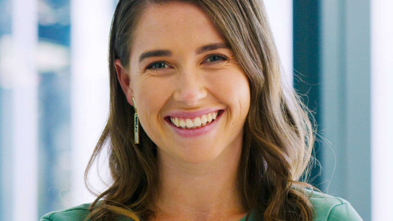 A woman with long brown hair and green eyes smiles warmly at the camera, wearing a green top and rectangular earrings. The background is softly blurred with hints of light and blue.