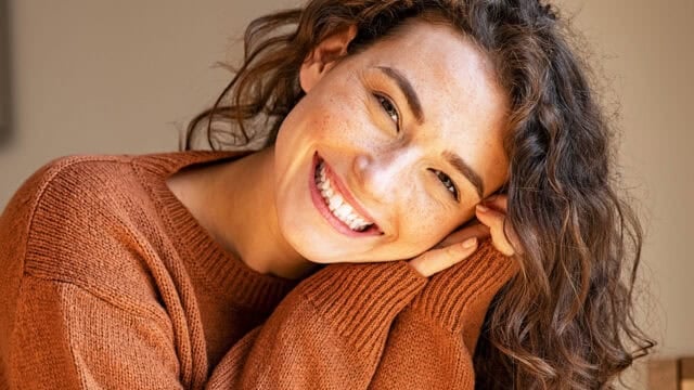 A person with curly brown hair, wearing a rust-colored sweater, smiles warmly at the camera with their head resting on their hand.