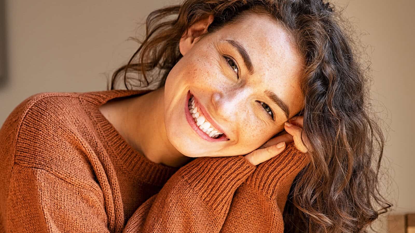 A person with curly brown hair, wearing a rust-colored sweater, smiles warmly at the camera with their head resting on their hand.