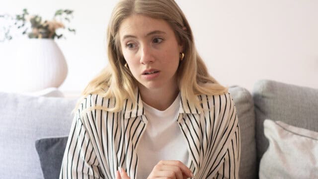 A woman with long blonde hair sits on a couch wearing a white shirt with black stripes, looking concerned or worried. A plant in a white vase is blurred in the background.