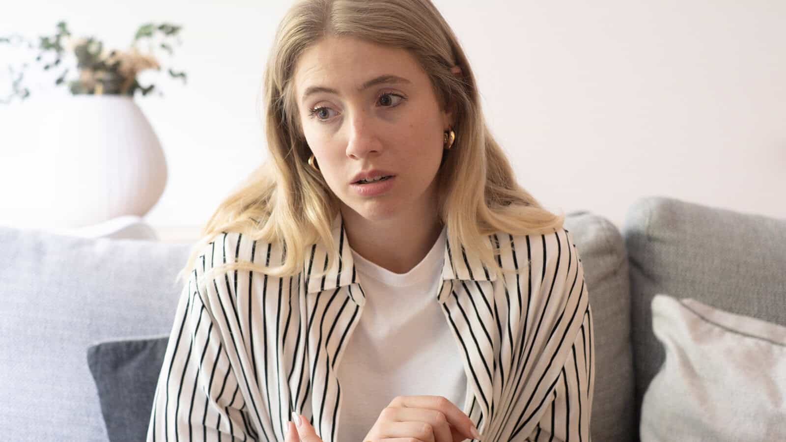 A woman with long blonde hair sits on a couch wearing a white shirt with black stripes, looking concerned or worried. A plant in a white vase is blurred in the background.
