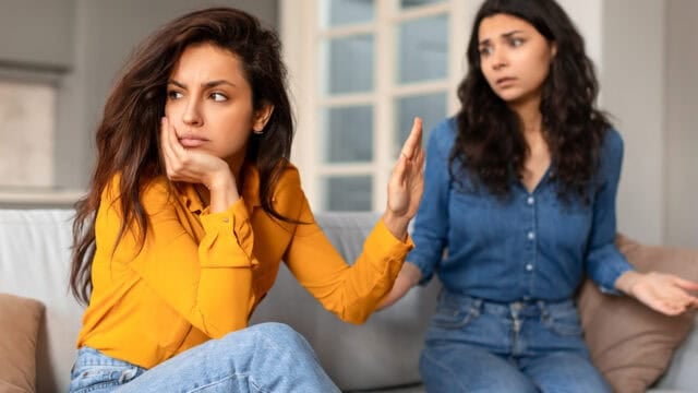 Two women sit on a couch; the woman in front looks upset and gestures to stop, while the woman behind her appears concerned and confused.