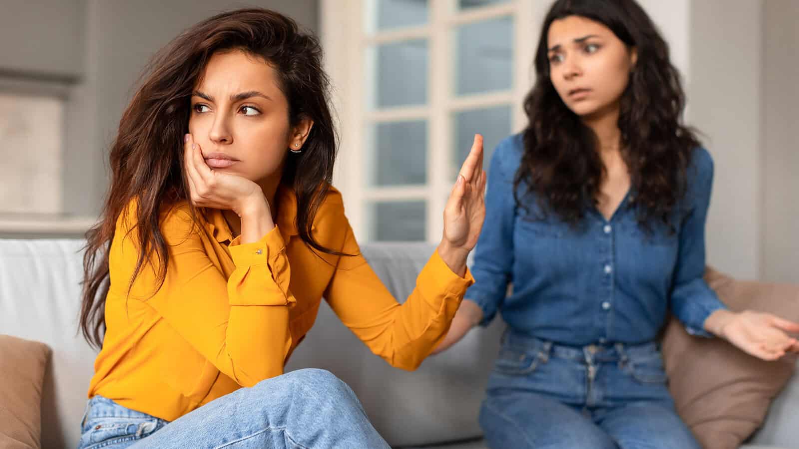 Two women sit on a couch; the woman in front looks upset and gestures to stop, while the woman behind her appears concerned and confused.