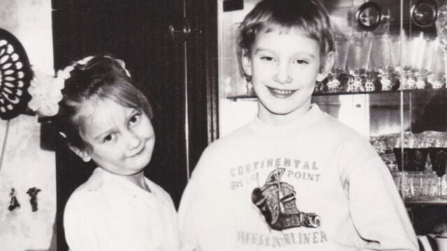 Two young children smiling and standing close together indoors, in front of a glass cabinet. The photo is black and white, capturing a sense of nostalgia from before smartphones—when tiny freedoms filled childhood, and happiness was simply found in each other's company.