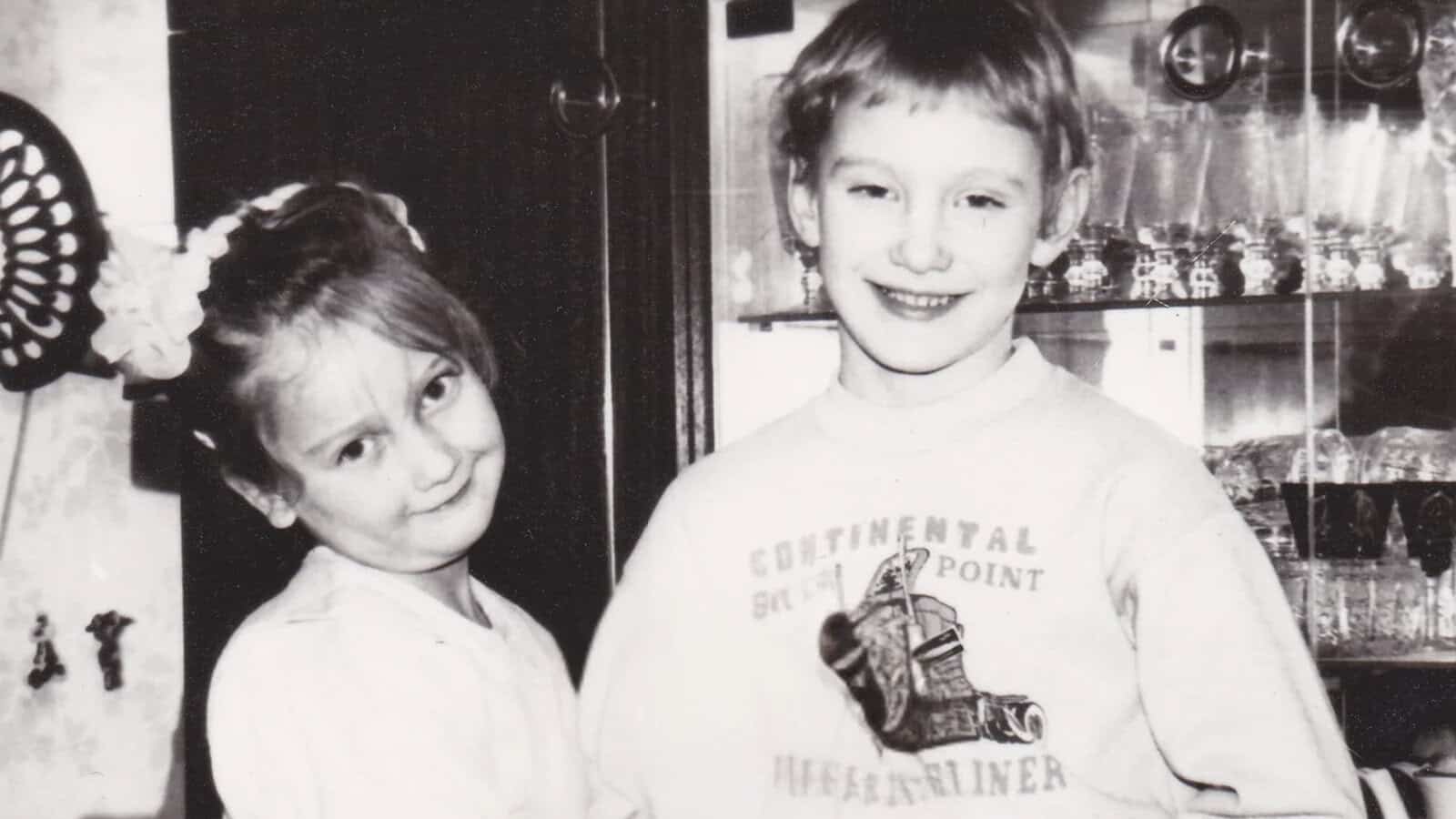 Two young children smiling and standing close together indoors, in front of a glass cabinet. The photo is black and white, capturing a sense of nostalgia from before smartphones—when tiny freedoms filled childhood, and happiness was simply found in each other's company.