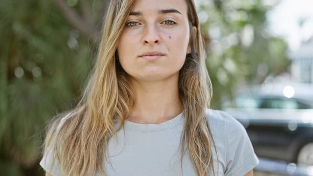 A young woman with long blonde hair and a neutral expression stands outdoors, wearing a light gray t-shirt. Greenery and blurred cars appear in the background.