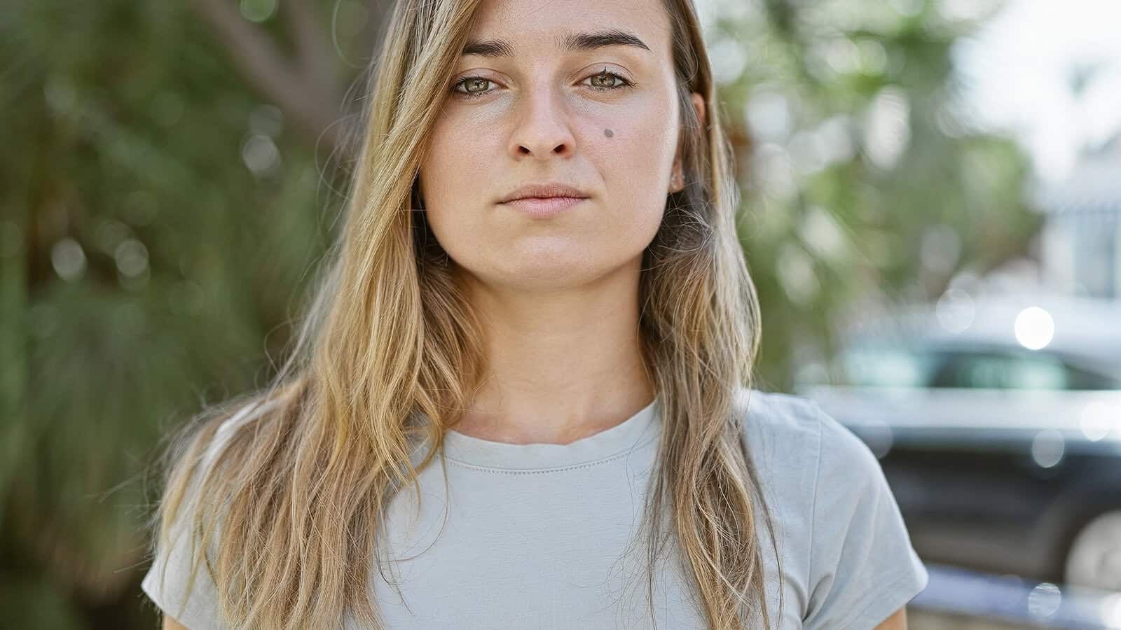 A young woman with long blonde hair and a neutral expression stands outdoors, wearing a light gray t-shirt. Greenery and blurred cars appear in the background.