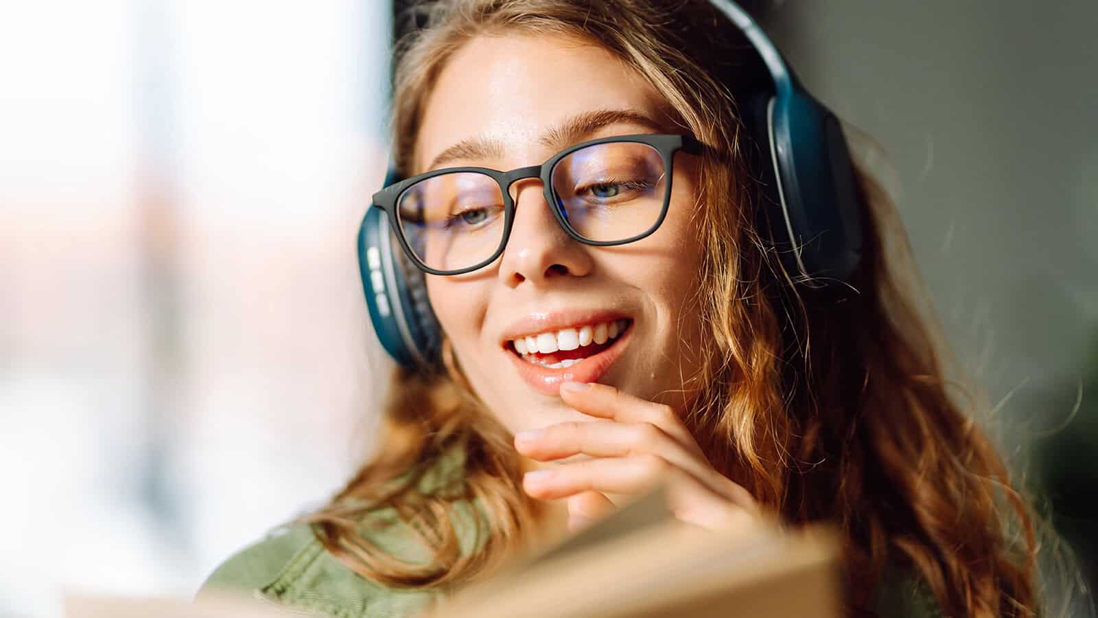 A young woman with glasses and headphones smiles while reading a book, holding her hand near her mouth in a relaxed, thoughtful pose.