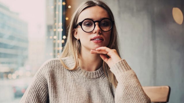 A woman with long blonde hair and round glasses sits indoors by a window, resting her chin on her hand and wearing a beige knit sweater. Blurred cityscape and string lights are visible in the background.