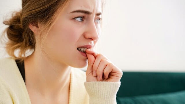 A young woman with light brown hair in a loose bun looks anxious, biting her fingernail while wearing a cream sweater. She gazes off to the side with a worried expression.