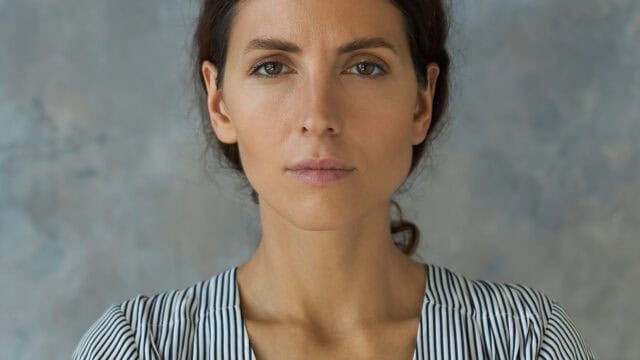 A woman with long brown hair in a striped shirt stares directly at the camera with a neutral expression, standing in front of a gray-blue mottled background.