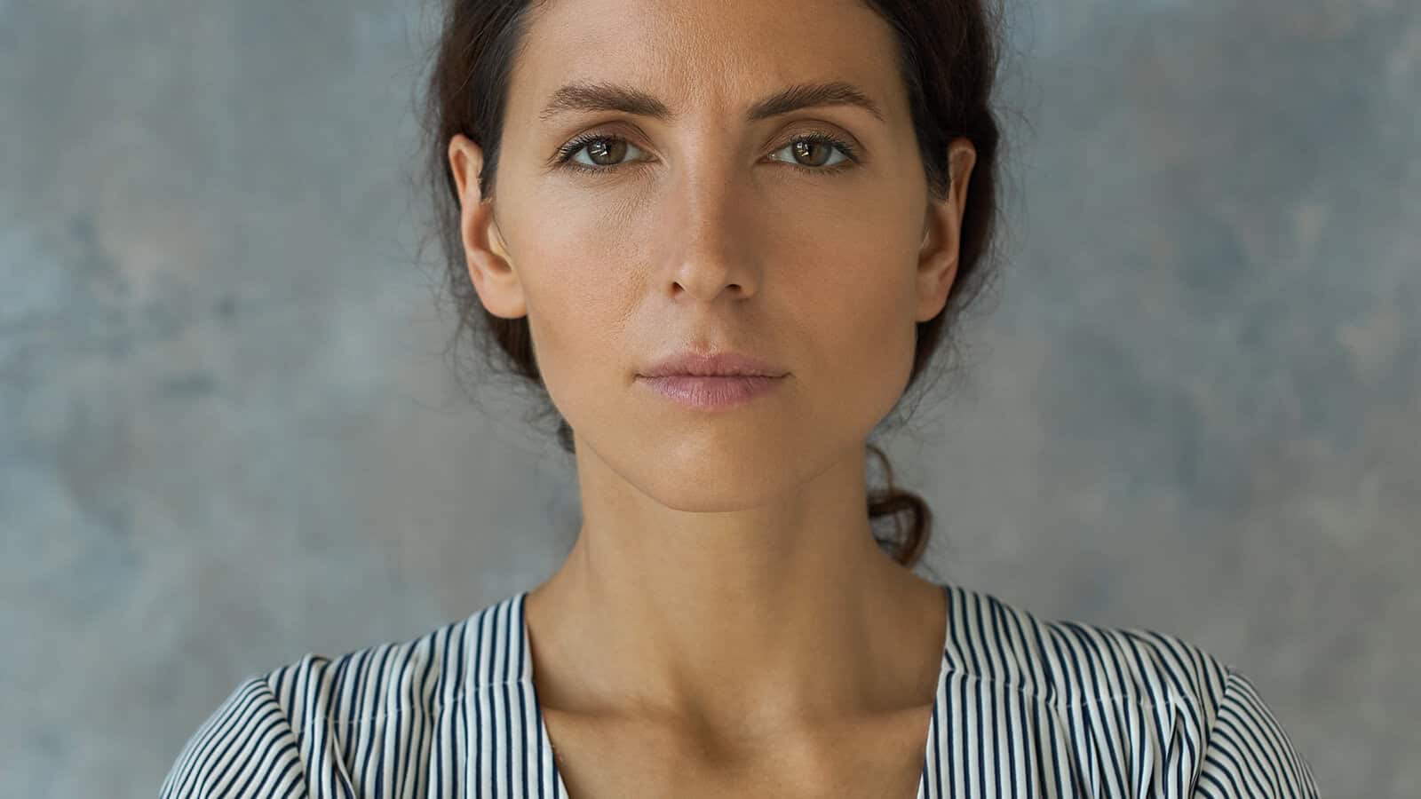 A woman with long brown hair in a striped shirt stares directly at the camera with a neutral expression, standing in front of a gray-blue mottled background.