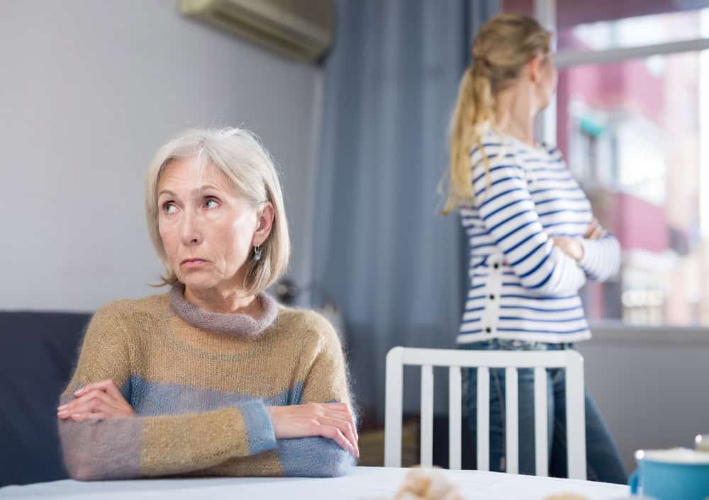 An older woman sits at a table with arms crossed, looking upset, perhaps reflecting on the behaviors to avoid as relationships change with getting older. A younger woman stands in the background, also with arms crossed, facing away and looking out the window—an image that speaks to the complexities of adult children respect within families.