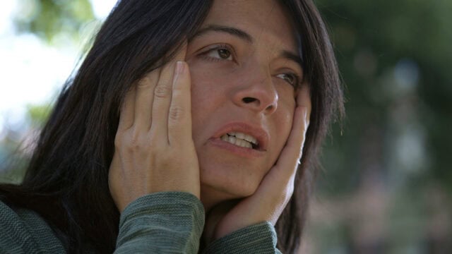 A woman with long dark hair holds her face with both hands, looking distressed or worried, outdoors with a blurred green background.
