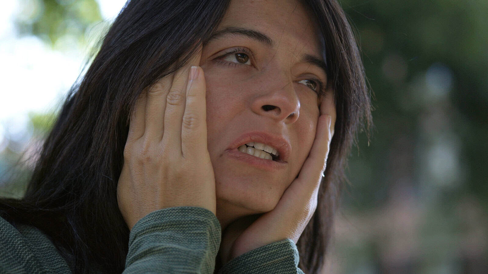 A woman with long dark hair holds her face with both hands, looking distressed or worried, outdoors with a blurred green background.