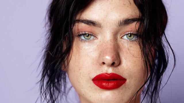 A person with light green eyes, prominent freckles, natural makeup, and bright red lipstick looks directly at the camera. Their dark hair is loosely framing their face against a pale purple background.
