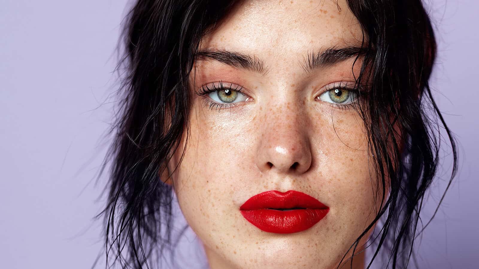 A person with light green eyes, prominent freckles, natural makeup, and bright red lipstick looks directly at the camera. Their dark hair is loosely framing their face against a pale purple background.